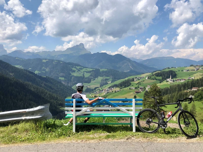 Gravelbike Holidays © Hotel Ostaria Posta Radfahrer auf Bank blickt auf grüne Berge und Dorf im Tal