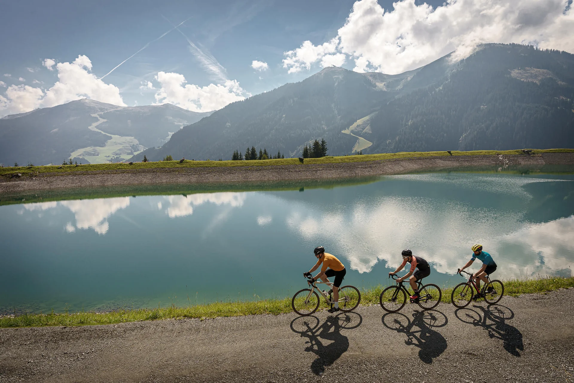 Gravelbike Holidays © Björn Hänssler Drei Radfahrer fahren an einem Bergsee mit Bergen und Wolken im Hintergrund