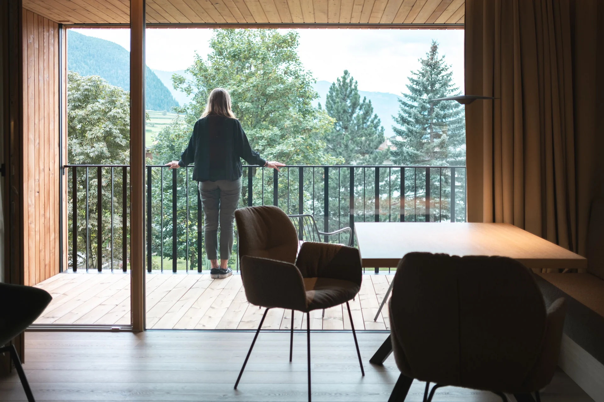 Gravelbike Holidays © Lunah Photography - Lukas Haberer & Hannah Beikircher Person blickt vom Balkon eines Holzhauses auf grüne Bäume und Berge
