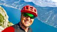 Gravelbike Holidays © Andrea Maino Mann mit rotem Fahrradhelm macht Selfie vor Berg und See