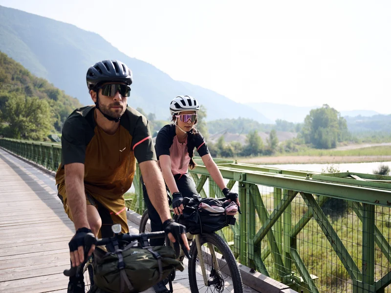 Gravelbike Holidays © Manuel Ferrigato Zwei Radfahrer auf einer Brücke vor bewaldeten Hügeln und Flusslandschaft