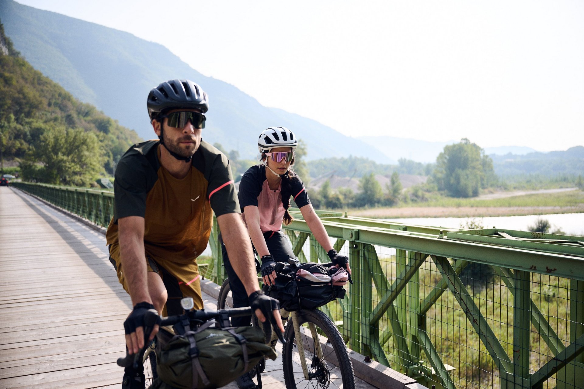 Gravelbike Holidays © Manuel Ferrigato Zwei Radfahrer auf einer Brücke vor bewaldeten Hügeln und Flusslandschaft