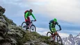 Gravelbike Holidays © Markus Greber Zwei Mountainbiker fahren an einem steilen Berg mit schneebedeckten Gipfeln im Hintergrund