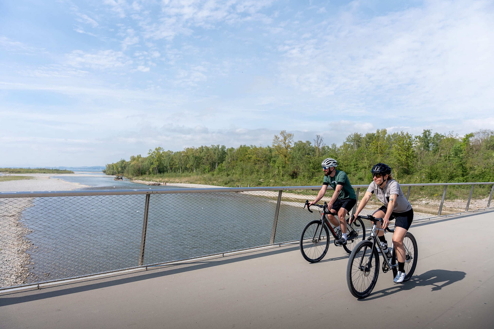 Gravelbike Holidays © Chris Gollhofer - Vorarlberg Tourismus Zwei Radfahrer auf einer Brücke neben einem Fluss unter bewölktem Himmel